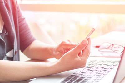 Midsection of woman using mobile phone by laptop at desk