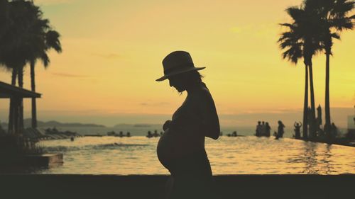 Pregnant woman standing at beach against sky during sunset