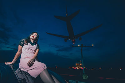 Young woman sitting outdoors while airplane flying against sky at night