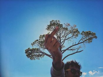 Low angle view of man showing middle finger with tree in background against blue sky