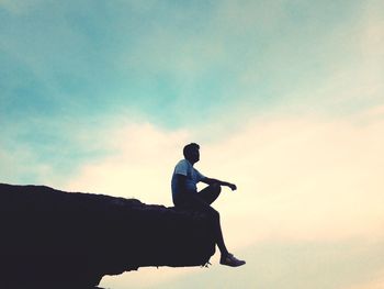 Low angle view of man sitting on cliff against sky