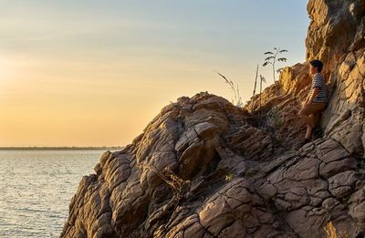 A teenage boy standing on rock by sea against sky