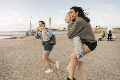 Female friend giving piggyback ride while teenage girl photographing at beach