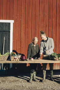 Full length of male and female farmers discussing over vegetables at table against barn