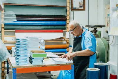 Side view of concentrated senior male artisan with white hair in apron and eyeglasses standing at table with pile of papers and notebook in printing workshop