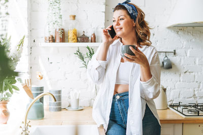 Attractive young woman plus size body positive in white shirt talking on cellphone at home kitchen