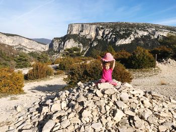 Girl holding hat while sitting on heap on rocks against rock formations