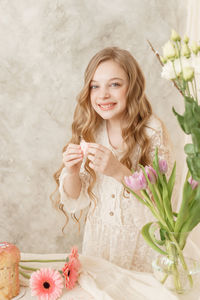 A girl with long hair in a light dress is sitting at the easter table with cakes, spring flowers 