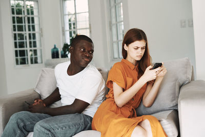 Sad man and woman using phone while sitting on sofa