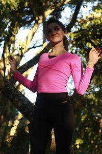 Low angle view of woman looking away with arms raised while standing against trees