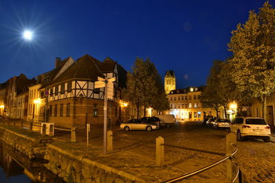 Illuminated street by buildings against sky at night