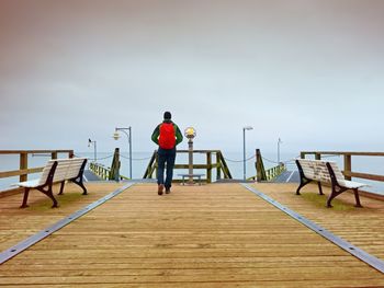 Tourist with red backpack on wooden pier above sea. man in trekking suit in harbor in rainy day.