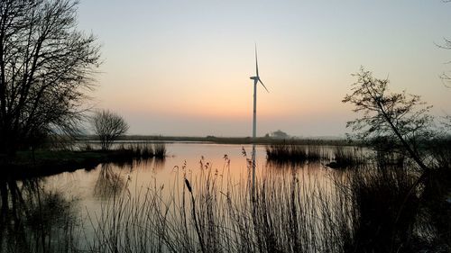 Wind turbine by lake against sky during sunset