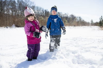 Playful siblings enjoying on snow covered field against sky