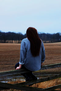 Rear view of woman sitting on railing at farm field against sky