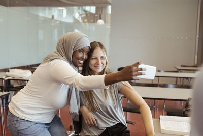 Smiling woman in hijab taking selfie with friend on smart phone at classroom