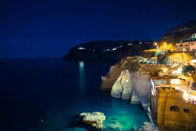 Illuminated buildings by sea against clear blue sky at night