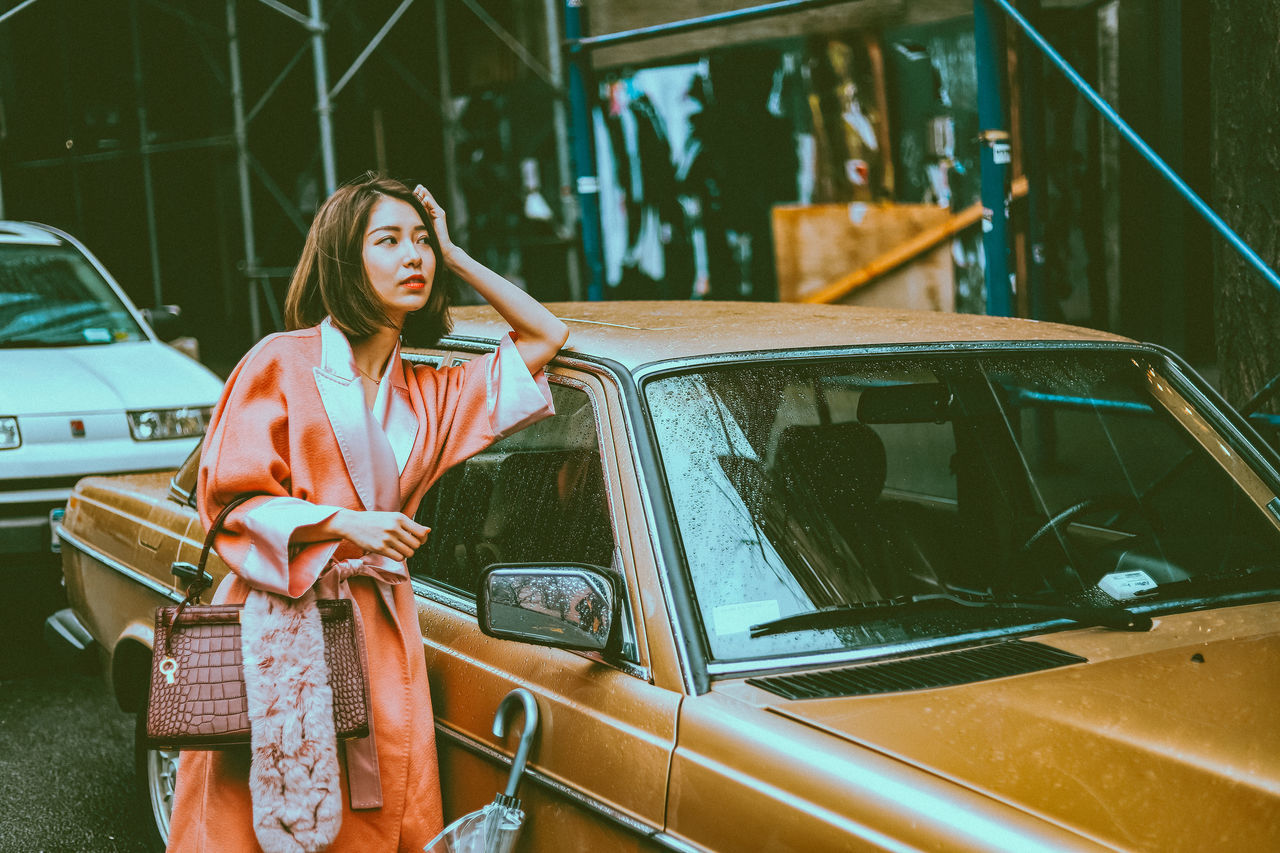 car, mode of transportation, motor vehicle, transportation, one person, adult, women, vehicle, young adult, lifestyles, land vehicle, standing, fashion, female, portrait, architecture, retro styled, travel, emotion, looking, front view, casual clothing, clothing, hairstyle, three quarter length, outdoors