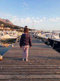 Rear view of girl walking on jetty amidst boats at harbor against sky