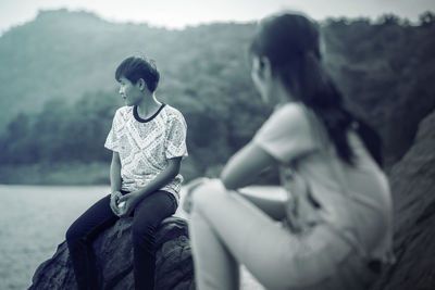 Teenage girl looking at boy sitting on rock by sea