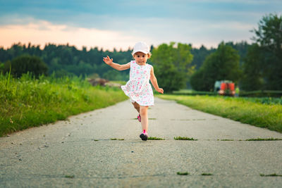 Baby girl with cap outdoors running on a rural road leading from forest.