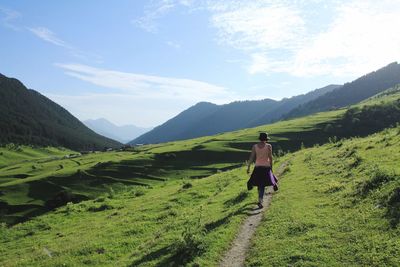 Rear view of woman walking on footpath amidst grassy field against cloudy sky