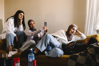 Young women talking with each other by friends taking selfie through mobile phone at home