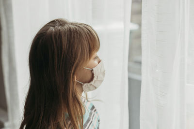 A sad dark-haired schoolgirl looks out the window wearing a mask. self-isolation period. patient 