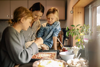 Mother and son watching boy spreading cheese on pizza while standing in kitchen at home