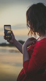 Side view of woman photographing with mobile phone at beach during sunset