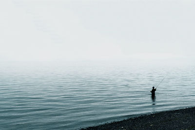 Fisherman casting fly line off shore with dense fog over water.