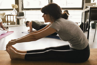 Full length side view of flexible mature businesswoman touching toes while practicing yoga on mat at workplace