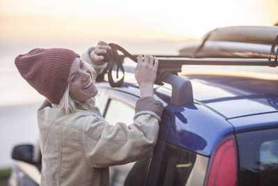 Happy young woman wearing warm clothes fastening surfboard on car roof