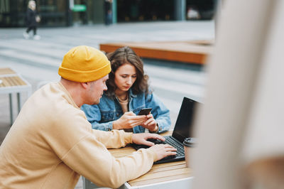 Stylish young couple freelancers working on laptop in street cafe, woman using mobile