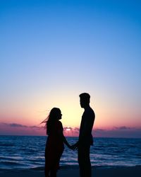 Silhouette couple holding hands at beach against sky during sunset