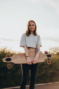 Portrait of smiling woman holding skateboard against sky