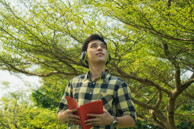 Low angle view of young man wearing headphones while holing book at park