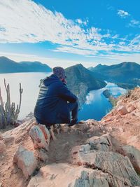 Rear view of man sitting on rock against sky