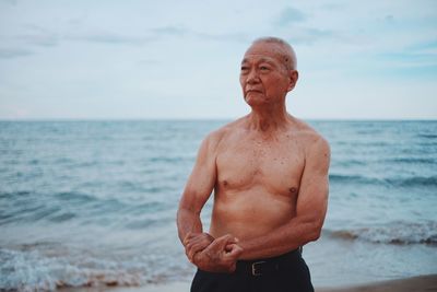 Shirtless senior man showing muscles while standing at beach against sky during sunset