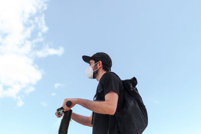 Low angle side view of anonymous male in respiratory mask and black clothes driving electric scooter under blue cloudy sky while looking forward