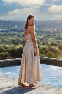 Young brunette woman standing next to a pool, wearing a cream white dress