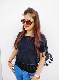 Beautiful young woman wearing sunglasses standing against white wall