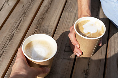 Hands of two people holding beige paper cups of cappuccino on wooden striped table outside, coffee