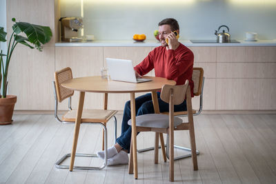 Smiling man working from home as freelancer using laptop, speaking on smartphone during work break