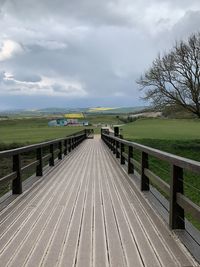 Narrow footbridge along trees on landscape against sky