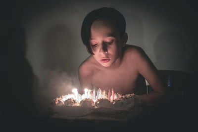 Shirtless boy blowing birthday candles on cake in dark