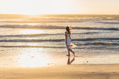 Side view of young woman walking at beach against clear sky during sunset