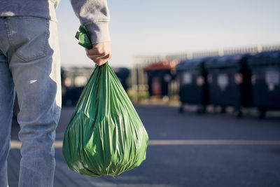 Man walking with rubbish. rear view of person carrying plastic bag against garbage cans on street.