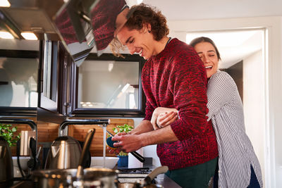 Side view delighted young female in casual clothes standing behind and embracing smiling boyfriend washing dishes in modern kitchen