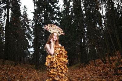 Thoughtful young woman covered with leaves standing at forest during autumn